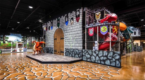 A colorful indoor play area featuring a castle structure, slides, and a dragon statue on a stone-patterned floor.