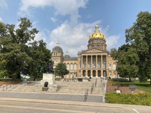 Iowa State Capitol building with a golden dome, surrounded by trees and a statue in front, under a partly cloudy sky.
