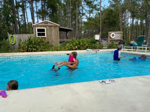 A child is being lifted in the water by an adult, while others swim nearby in a pool surrounded by trees.
