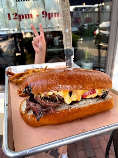 A close-up of a sandwich with meat and cheese, served on a tray with fries, and a hand making a peace sign in the background.