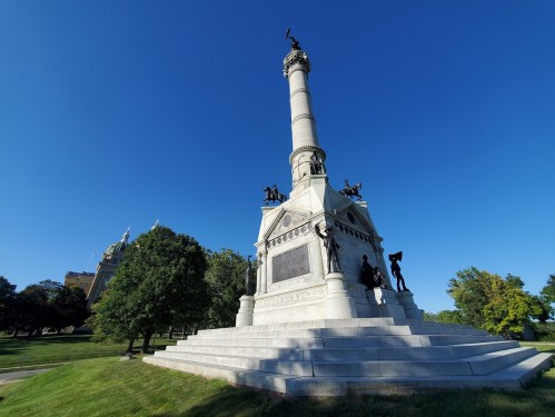 A tall monument with a column and statues, set against a clear blue sky and surrounded by trees.