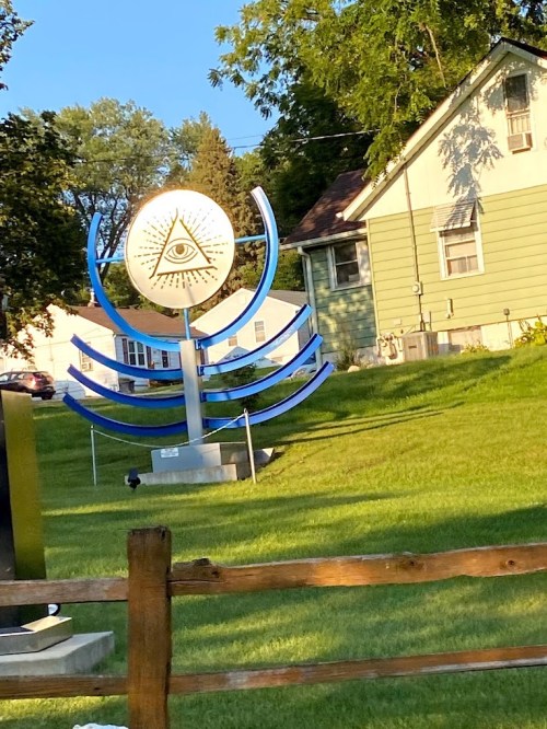 A large, stylized sculpture featuring an eye symbol, set in a grassy area near a house.