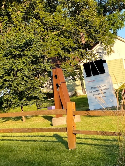 A large wooden clip and sign in a park reminding visitors to keep the area clean, surrounded by greenery.