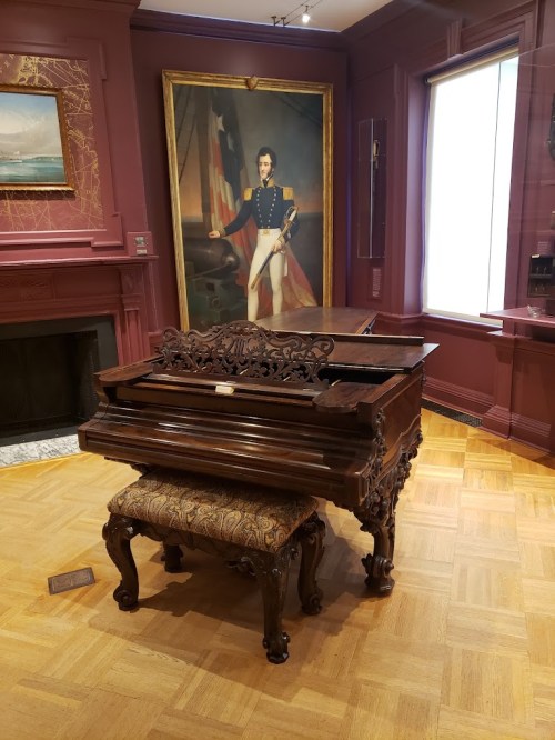 A vintage piano with ornate details in a room featuring a large portrait of a man in military attire.
