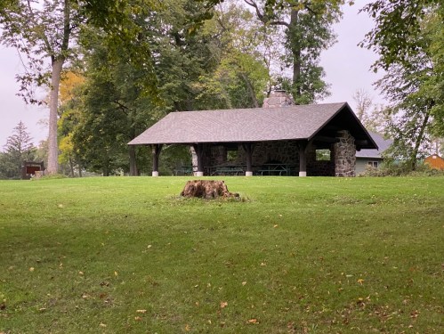 A stone pavilion with a sloped roof surrounded by trees, set on a grassy area with a tree stump in the foreground.