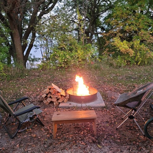 A cozy campfire surrounded by chairs and firewood, set in a wooded area with autumn foliage.