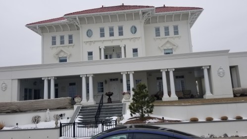 A large, elegant white mansion with a red roof, featuring columns and a wide staircase, set in a snowy landscape.