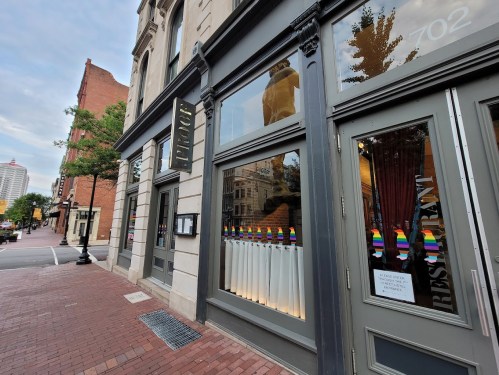 Exterior of a restaurant with rainbow flags in the windows, located on a brick street in an urban area.