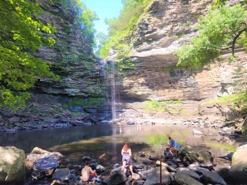A serene waterfall cascades into a pool surrounded by rocky cliffs and lush greenery, with people enjoying the scene.