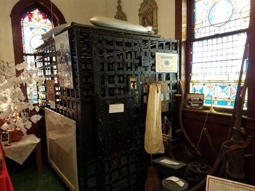 A large, historic jail cell displayed in a museum, surrounded by artifacts and stained glass windows.