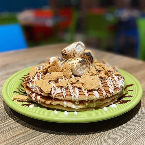 A stack of pancakes topped with toasted marshmallows, graham cracker pieces, and drizzled with chocolate sauce on a green plate.