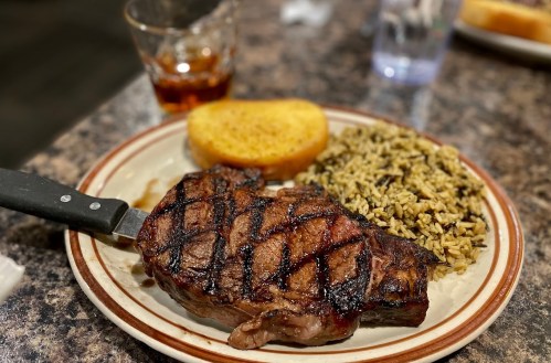 A grilled steak with grill marks, a slice of garlic bread, and a serving of rice on a plate, with a drink in the background.