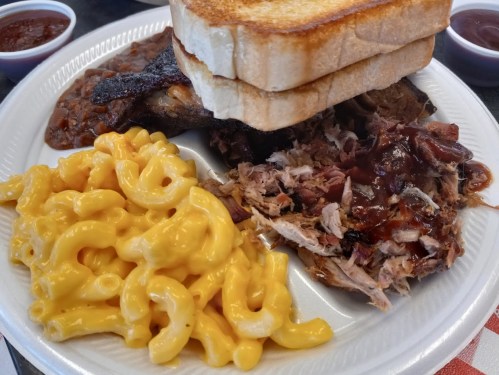 A plate of barbecue featuring pulled pork, macaroni and cheese, baked beans, and toasted bread, with barbecue sauce on the side.