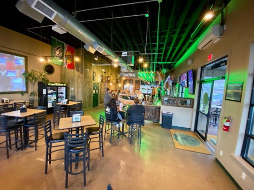 Interior of a casual bar with tables, a bar area, and screens showing sports, featuring green lighting and decor.