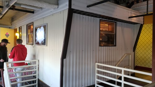 A small, modern building with a corrugated metal exterior and large windows, featuring a staircase and outdoor seating area.