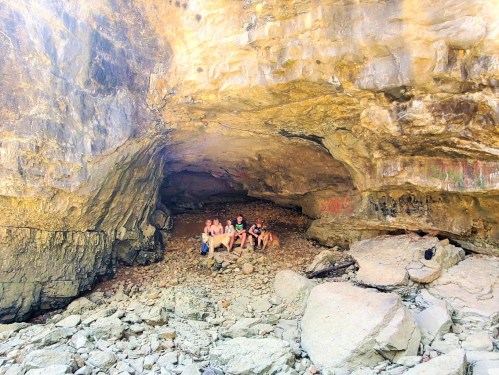 Four people sitting on rocky ground inside a large cave, with textured stone walls and faint markings in the background.