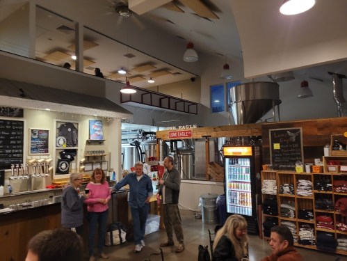 A cozy brewery interior with patrons chatting, a bar with taps, and brewing equipment in the background.