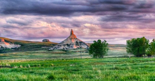 A dramatic landscape featuring a prominent rock formation, green fields, and a cloudy sky in Nebraska.