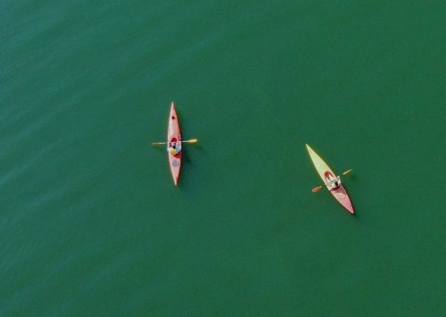 Two kayakers paddle on a calm, greenish-blue body of water, one in a red kayak and the other in a yellow kayak.