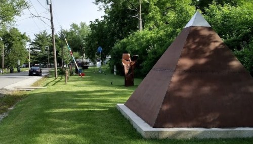 A pyramid sculpture on a grassy area beside a road, with trees and other abstract sculptures in the background.