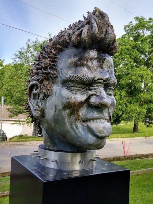 A large, expressive sculpture of a face sticking out its tongue, with spiked collar and textured hair, set outdoors.