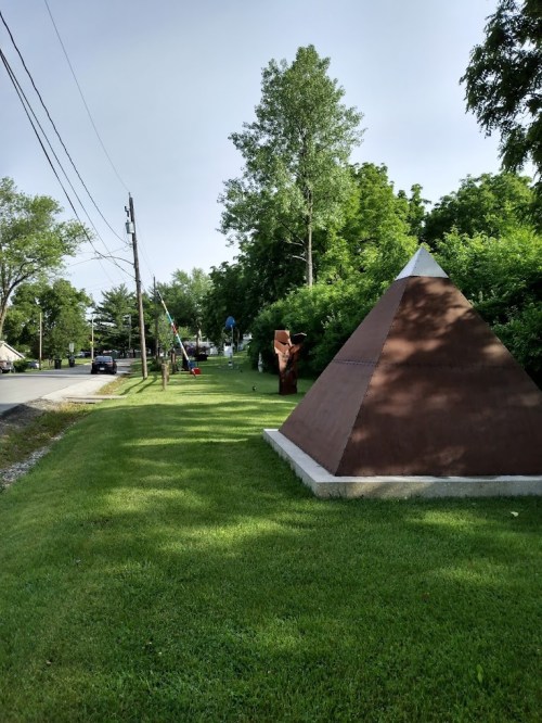 A pyramid-shaped sculpture on a grassy area beside a road, with trees and a few people in the background.