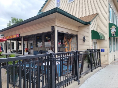 A restaurant exterior with outdoor seating, featuring a stone wall and large windows, under a cloudy sky.