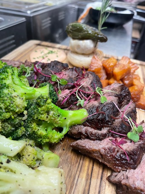 A wooden platter featuring grilled steak, broccoli, sweet potatoes, and a decorative garnish.
