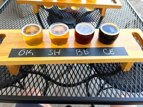 A wooden flight board with four small glasses of beer, labeled "OK," "SH," "BB," and "CE," on a black table.