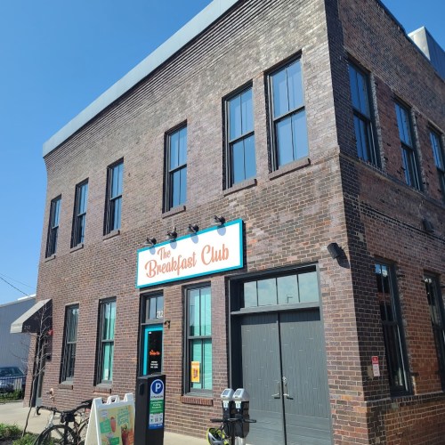 Exterior of a brick building with a sign reading "The Breakfast Club" and a bike parked outside. Clear blue sky.