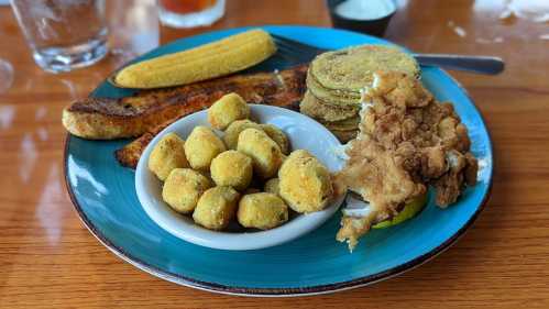 A blue plate with fried chicken, fried green tomatoes, corn on the cob, and a bowl of fried okra.