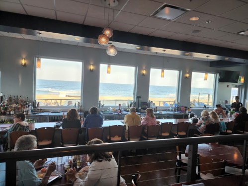 A restaurant interior with patrons at the bar, large windows showcasing a beach view and ocean waves outside.