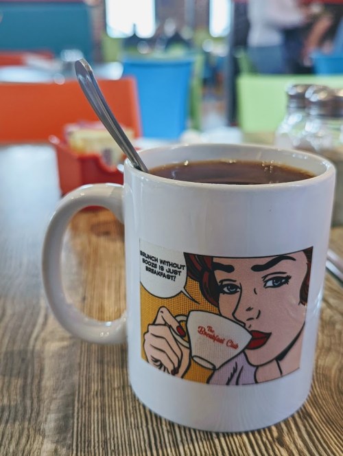 A coffee mug with a retro illustration and text about brunch, placed on a wooden table in a colorful café.