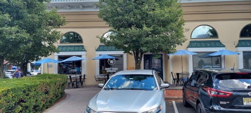 A view of a café with outdoor seating under blue umbrellas, surrounded by trees and parked cars.