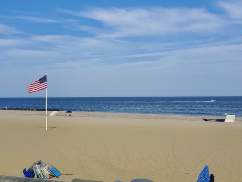 A sandy beach with an American flag, calm ocean waters, and a boat in the distance under a blue sky.