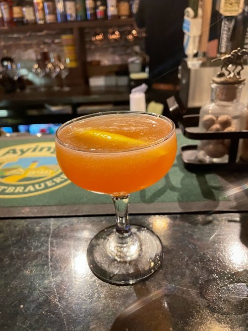 A vibrant orange cocktail with a twist of lemon served in a classic coupe glass on a bar counter.