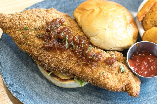 A plate featuring a crispy fried fish fillet with sauce on a bun, accompanied by potato wedges and a small cup of ketchup.