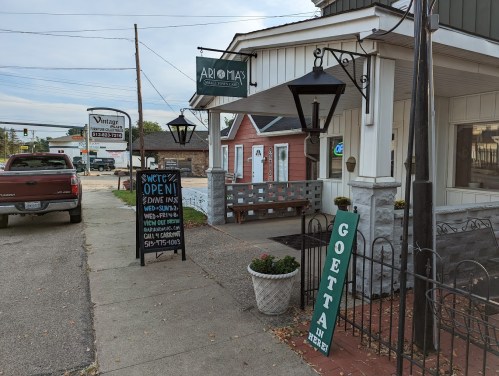 A street view of a restaurant with a sign saying "Open," surrounded by a sidewalk, plants, and parked vehicles.