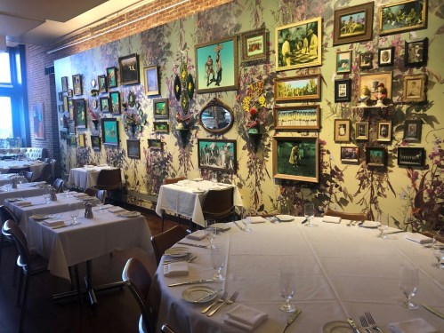 A restaurant interior featuring elegantly set tables and a vibrant wall adorned with framed art and floral wallpaper.