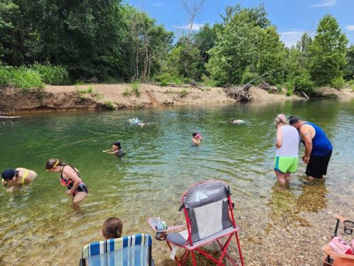 A group of people enjoying a sunny day by a clear river, some wading in the water and others sitting on the shore.