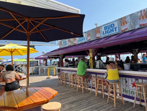 A vibrant beach bar scene with patrons at the counter, colorful umbrellas, and a sunny sky in the background.