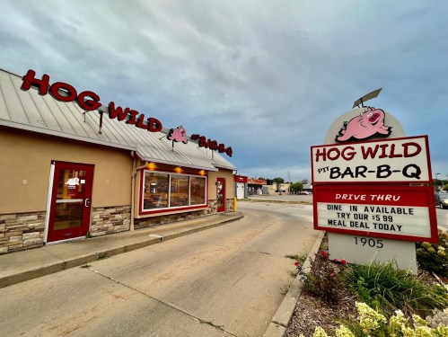 Exterior of Hog Wild Bar-B-Q restaurant with a sign advertising drive-thru and meal deals. Cloudy sky in the background.