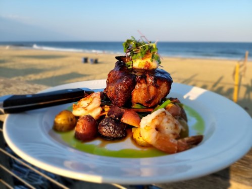 A gourmet dish featuring grilled shrimp, potatoes, and a meat patty, served on a beachside table with ocean views.