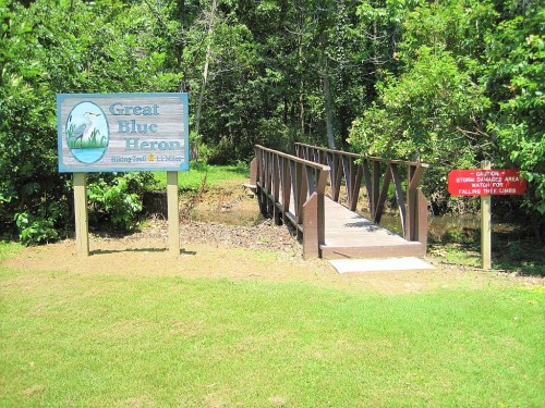 Sign for Great Blue Heron with a wooden bridge over a small waterway, surrounded by greenery. Caution sign nearby.