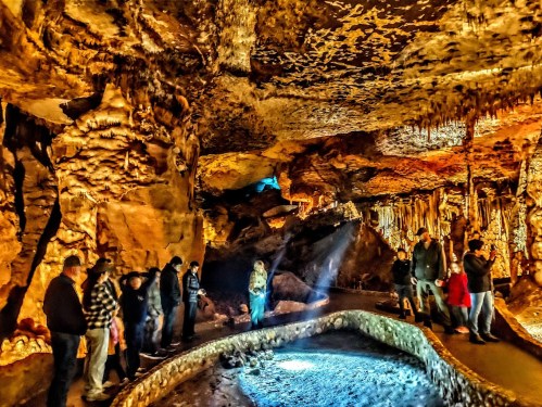 Visitors explore a stunning cave with illuminated rock formations and a shimmering pool of water.