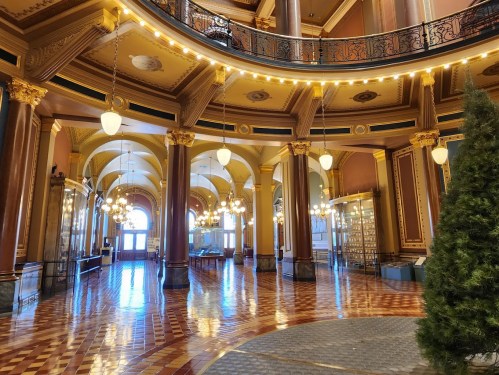 Interior of a grand hall with ornate columns, decorative lighting, and a polished floor, featuring large windows.