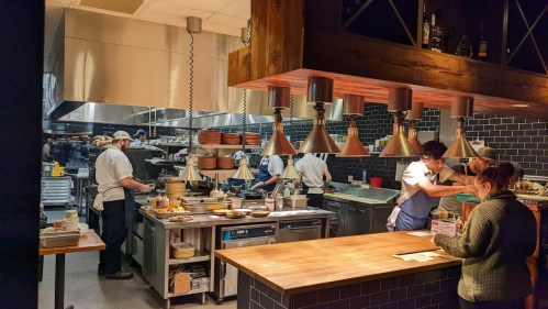A busy restaurant kitchen with chefs preparing food and a customer at the counter, surrounded by cooking equipment.