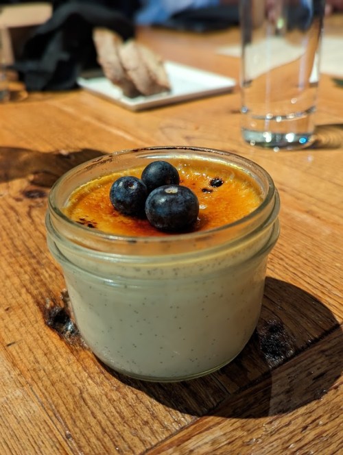 A creamy dessert in a jar topped with blueberries and a caramelized surface, placed on a wooden table.