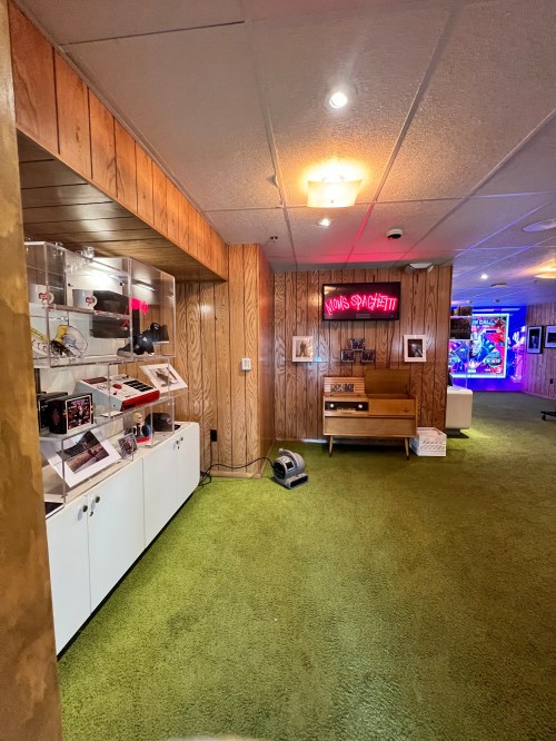 A retro-themed game room with wooden paneling, green carpet, and vintage gaming consoles displayed in glass cases.