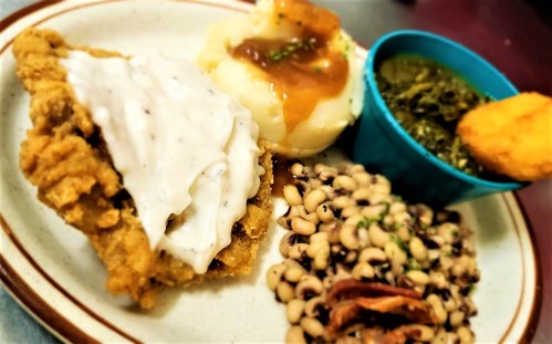 A plate featuring fried chicken with gravy, mashed potatoes, collard greens, black-eyed peas, and a cornbread muffin.